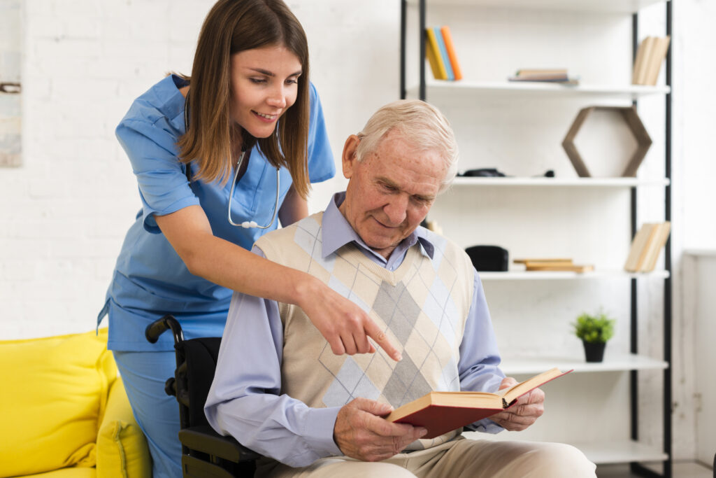 a woman in a blue scrub outfit leaning over a mans back pointing as the man in the chair is reading a book in a blue and grey sweater.